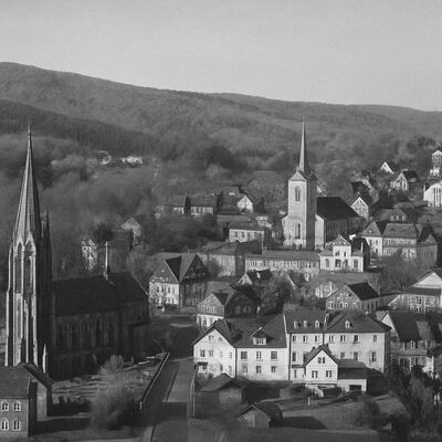 Historische Fotografie vom Stadtkern der Stadt Kirchen Schwarz-weiß-Fotografie des historischen Stadtkerns der Stadt Kirchen mit zwei Kirchen und traditionellen Fachwerkhäusern, eingebettet in eine hügelige Waldlandschaft.