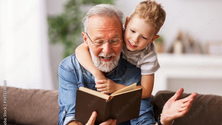 Senior sitzt auf einer Couch, hinter ihm sitzt ein kleiner Junge. Beide lesen freudig in einem Buch.