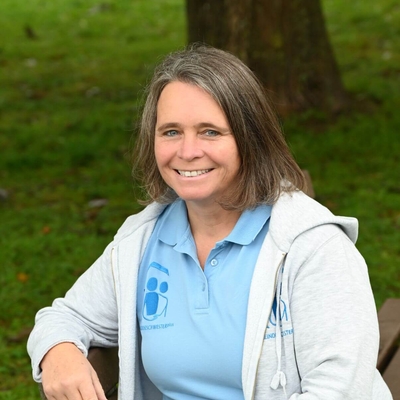 Lächelnde Frau sitzt auf einer Holzbank im Grünen und blickt in die Kamera.